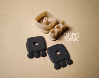 May include: Two black polymer clay earrings with a square shape and three small round accents hanging from the bottom. The earrings are shown next to their molds, which are beige and made of plastic.