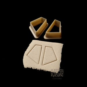 May include: Two beige plastic cookie cutters shaped like diamonds, with a beige clay surface showing the imprint of the cutters. The clay surface has the text "HARPER BAYNE CUTTERS" printed on it.