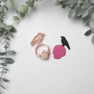 May include: Halloween-themed cookie cutters featuring a light brown bird, a light brown skull, and a pink skull with a black bird. The cutters are displayed on a white surface, with green foliage in the background.