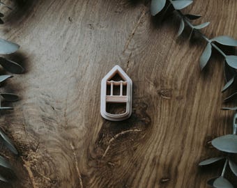 May include: A light pink cookie cutter in the shape of a house, with window and door details. The cutter is centered on a dark wood surface, surrounded by green leafy branches.