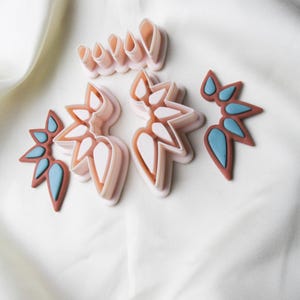 May include: A selection of pink and brown floral-shaped cookie cutters with blue accents. The cutters are arranged on a white fabric background. The shapes include petal and leaf designs, ideal for baking and crafting.