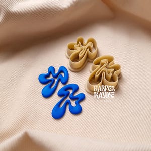 Op de afbeelding: Twee blauwe plastic koekjesvormen in de vorm van abstracte swirls. De vormen liggen op een beige stoffen achtergrond. De tekst "HARPER BAYNE CUTLERS" is zichtbaar in de afbeelding.