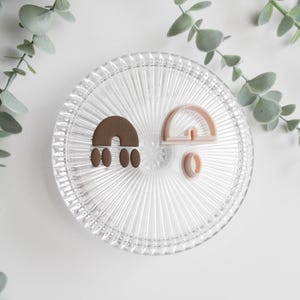 May include: Two cookie cutters in the shape of rainbows and a circle are displayed on a clear, ridged glass plate. One rainbow is brown, the other is pink. The plate is on a white surface, with green leafy sprigs in the background.