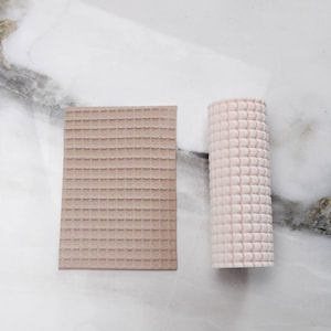 Può includere: Due stampi in silicone strutturati. Uno è rettangolare, color beige, e presenta un motivo a griglia di piccoli quadrati. L'altro è cilindrico, rosa chiaro, e ha un motivo a griglia simile. Entrambi sono su una superficie di marmo bianco e grigio.