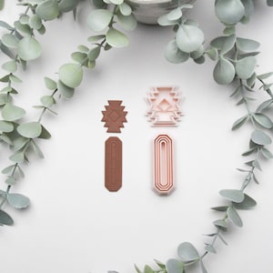 Könnte beinhalten: Zwei Sets von Ausstechern mit geometrischen Designs. Ein Set ist hellbraun, das andere hellrosa. Die Ausstecher sind auf einer weißen Oberfläche mit grünen Eukalyptusblättern und einem Topf im Hintergrund angeordnet.