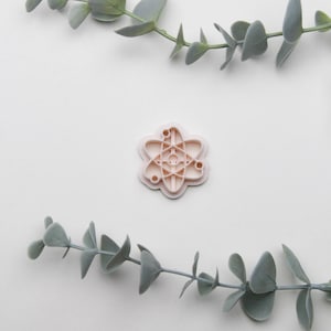 May include: A pink 3D printed atom symbol on a white background with eucalyptus branches on either side.