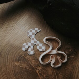 May include: Two abstract-shaped cookie cutters. One is a light pink plastic cutter, and the other is a matching shape with a tan and white checkered pattern. The cutters are on a wooden surface.