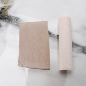 May include: A beige, rectangular fabric piece and a rolled-up cylinder of similar material. Both items have a textured, woven appearance. The items are displayed on a white and gray marble surface, creating a neutral and minimalist aesthetic.
