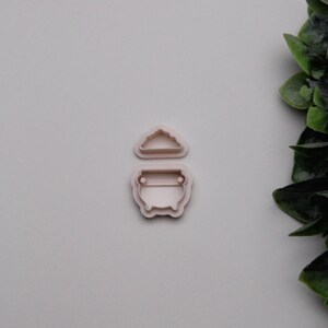 May include: Two light pink plastic cookie cutters. One is shaped like a cauldron with a handle, and the other is a cloud. The cookie cutters are on a light grey background with green leaves in the corner.