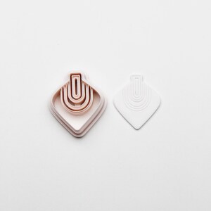 May include: A set of three light pink diamond-shaped cookie cutters with a layered design, featuring a curved, arched pattern. A separate white cookie cutter with a similar design is also present. The items are on a white background.