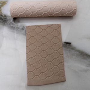 May include: A beige clay roller and a rectangular clay piece, both with a seashell pattern. The roller is cylindrical, and the clay piece is flat. The seashell pattern is embossed on both items, creating a textured surface.
