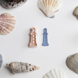 Puede incluir: Dos cortadores de galletas con forma de faro, uno marrón claro y otro azul, están rodeados de varias conchas marinas sobre un fondo blanco. Las conchas marinas varían en forma, tamaño y color, incluyendo marrón, blanco y beige.