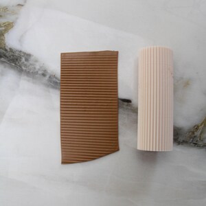 May include: A brown, rectangular piece with horizontal lines and a light pink, cylindrical object with vertical lines. Both objects are on a white and gray marble surface. The brown piece has a curved edge.