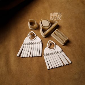 May include: A set of polymer clay earring cutters with the text "Harper Bayne Cutters" on the top. The cutters are beige and are used to create fringe earrings. Two pairs of white fringe earrings are shown on a brown suede background.