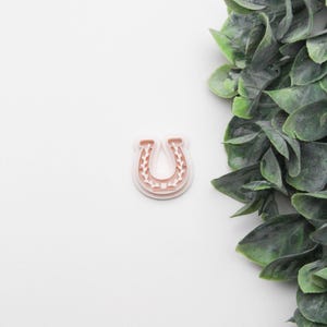 May include: A light pink horseshoe-shaped cookie cutter with a detailed floral design. The cutter is made of plastic and is set against a white background with green foliage in the upper right corner. This item is ideal for baking.