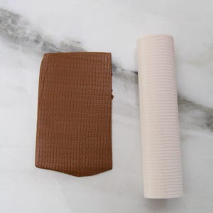May include: A rectangular piece of brown, textured material and a rolled, off-white, textured material are displayed on a white marble surface. The brown material has a knitted appearance. The off-white material is cylindrical.