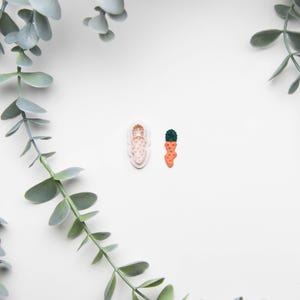 May include: A carrot-shaped cookie cutter and a miniature carrot decoration are displayed on a white surface, surrounded by green leafy sprigs. The cookie cutter is light pink, and the carrot decoration is orange with a green top.