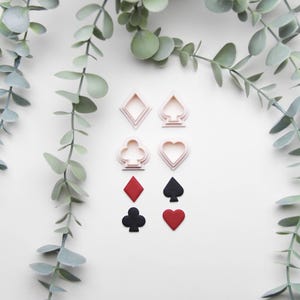 Könnte beinhalten: Ein Set mit sechs rosafarbenen Plastik-Ausstechern in Form von Spielkarten-Symbolen: Herzen, Karo, Pik und Kreuz. Außerdem gibt es drei Fondant-Ausschnitte: ein rotes Herz, ein schwarzes Pik und ein schwarzes Kreuz.