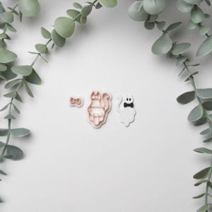 May include: Three Halloween-themed cookie cutters: a bow tie, a cat, and a ghost with a bow tie. The cat and bow tie cutters are light brown, while the ghost is white. Eucalyptus branches frame the top and sides on a white background.