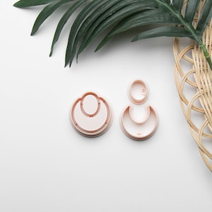 May include: Two pink plastic cookie cutters shaped like a crescent moon. The larger cutter has a smaller circle inside the crescent moon shape.