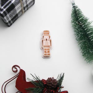 May include: A Christmas-themed flat lay. It features a light brown nutcracker cookie cutter, a miniature green Christmas tree, a red sled, and a wrapped gift with a black and white plaid pattern. The scene is set against a white background.