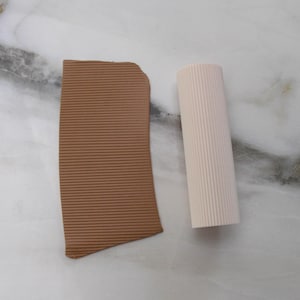 May include: A brown, rectangular piece of textured material next to a cylindrical, off-white object with vertical ridges. The objects are on a light-colored surface, possibly marble or stone.