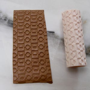 May include: Two textured clay tools. One is a rectangular slab in a brown hue, featuring a repeating pattern of squares and circles. The other is a cylindrical roller in a light pink color, with a similar embossed design. Both tools are for crafting.