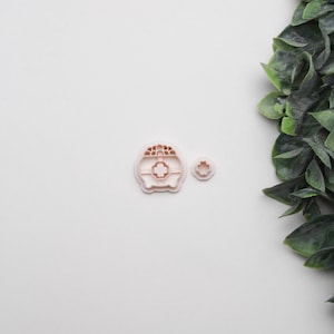 May include: Two light pink cookie cutters. One is a rounded shape with a cross in the centre and a grid pattern on top. The other is a smaller, flower-shaped cutter. The cutters are on a white surface.
