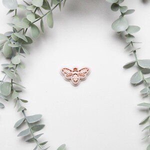 Peut inclure: Un emporte-pièce en forme d'abeille de couleur pêche clair. Le découpoir présente des motifs détaillés sur les ailes et le corps. L'image est posée sur un fond blanc, avec des feuilles d'eucalyptus vertes encadrant le haut et le bas.