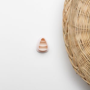 May include: A light pink, three-tiered candy corn shaped cookie cutter. The cutter is made of plastic and sits on a white surface. A woven basket is in the background.