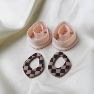 May include: Handmade polymer clay earrings with a black and tan checkerboard design. Two matching clay cutters are also visible. The earrings are oval-shaped with a central hole. The background is a white, silky fabric. The earrings are likely for pierced ears.
