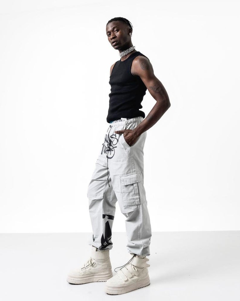 May include: A person wearing a black tank top, light gray cargo pants with black graphic print, and white high-top sneakers.