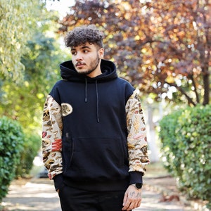 K&ouml;nnte beinhalten: Ein junger Mann tr&auml;gt einen schwarzen Hoodie mit einem bunten, gemusterten &Auml;rmel und schwarzen Hosen. Er steht in einem Park mit B&auml;umen im Hintergrund.