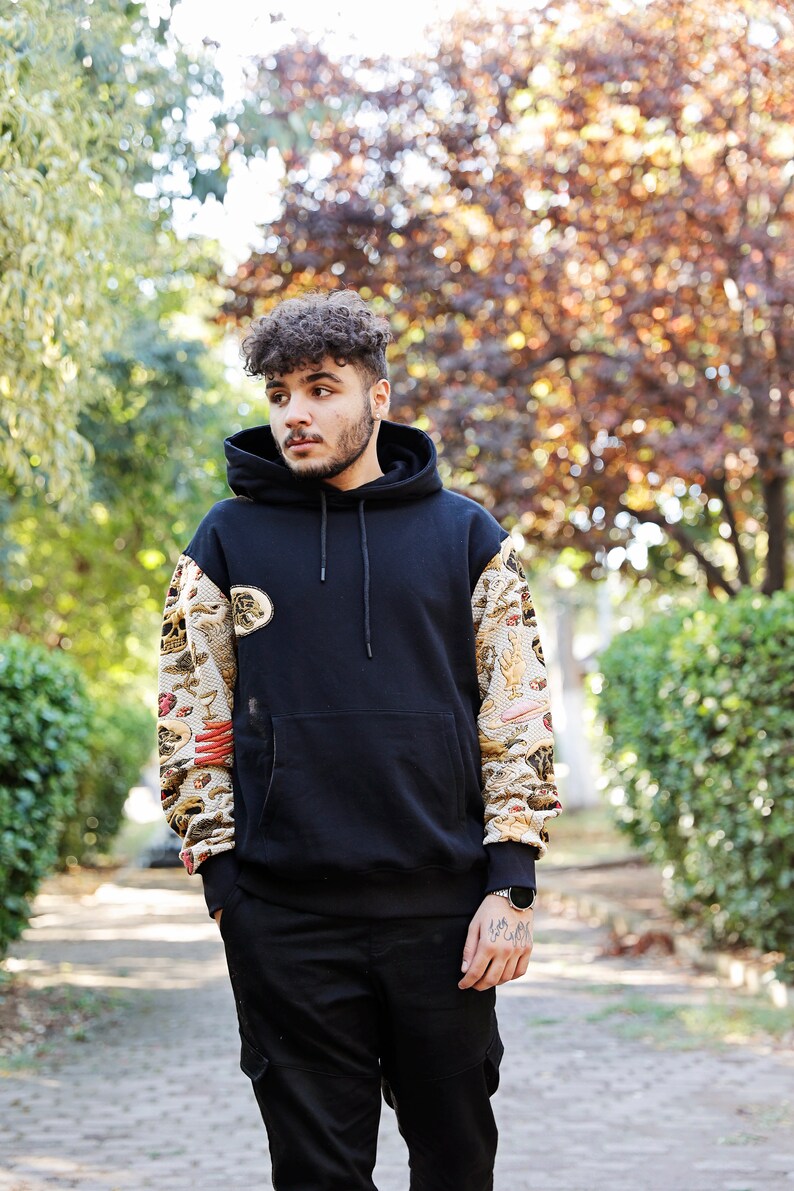 K&ouml;nnte beinhalten: Ein junger Mann tr&auml;gt einen schwarzen Hoodie mit einem bunten, gemusterten &Auml;rmel und schwarzen Hosen. Er steht in einem Park mit B&auml;umen im Hintergrund.