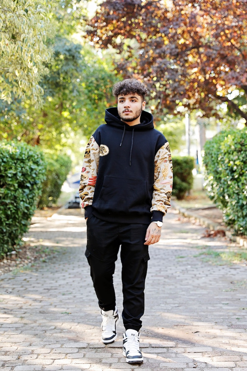K&ouml;nnte beinhalten: Ein Mann tr&auml;gt ein schwarzes Kapuzenpullover mit gemusterten &Auml;rmeln und eine schwarze Cargohose. Er tr&auml;gt wei&szlig;e Turnschuhe und eine Uhr.