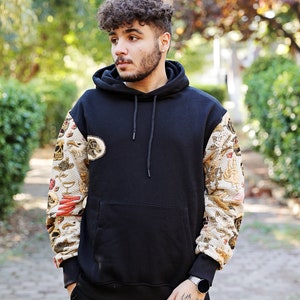 K&ouml;nnte beinhalten: Ein schwarzer Kapuzenpullover mit einem gemusterten Design an den &Auml;rmeln. Das Muster zeigt eine Vielzahl von Bildern, darunter Sch&auml;del, Blumen und andere Motive. Der Pullover hat eine K&auml;ngurutasche und eine Kapuze mit Kordelzug.