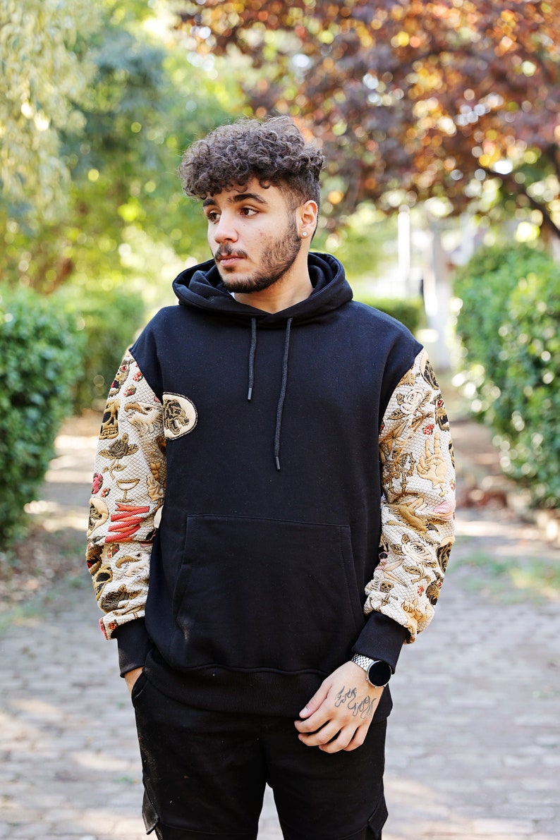 K&ouml;nnte beinhalten: Ein schwarzer Kapuzenpullover mit einem gemusterten Design an den &Auml;rmeln. Das Muster zeigt eine Vielzahl von Bildern, darunter Sch&auml;del, Blumen und andere Motive. Der Pullover hat eine K&auml;ngurutasche und eine Kapuze mit Kordelzug.