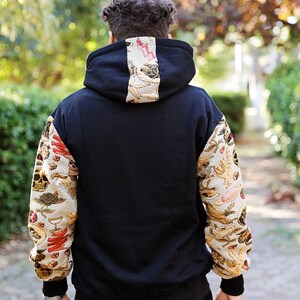 K&ouml;nnte beinhalten: Schwarzer Kapuzenpullover mit langen &Auml;rmeln, die mit einem gemusterten Design versehen sind. Das Muster ist eine Mischung aus Gold, Rot und Wei&szlig; mit verschiedenen Bildern.