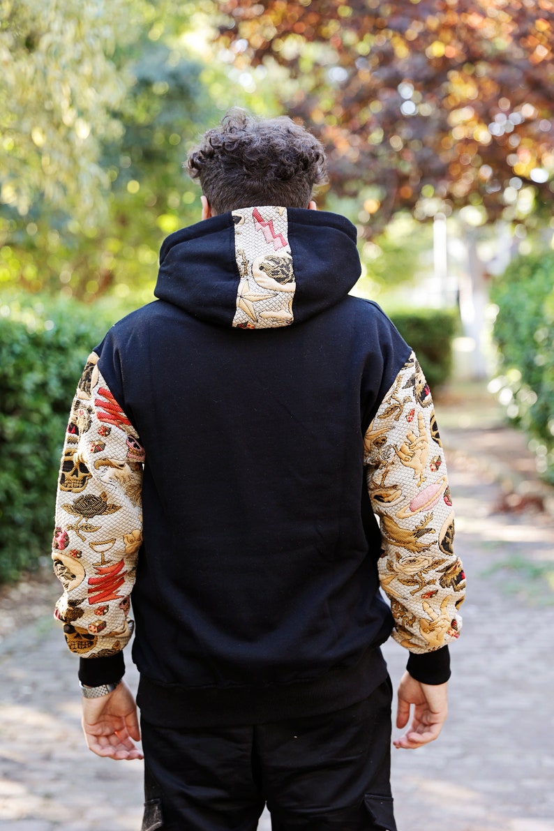 K&ouml;nnte beinhalten: Schwarzer Kapuzenpullover mit langen &Auml;rmeln, die mit einem gemusterten Design versehen sind. Das Muster ist eine Mischung aus Gold, Rot und Wei&szlig; mit verschiedenen Bildern.