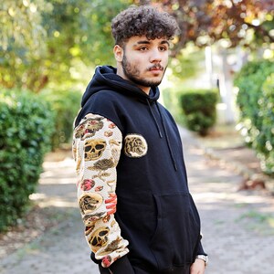 K&ouml;nnte beinhalten: Ein schwarzer Hoodie mit einem Totenkopf und anderen grafischen Designs auf dem linken &Auml;rmel. Der Hoodie hat eine gro&szlig;e Tasche auf der Vorderseite und eine Kapuze mit Kordelzug.
