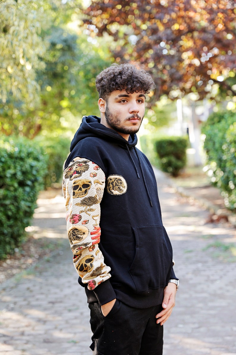 K&ouml;nnte beinhalten: Ein schwarzer Hoodie mit einem Totenkopf und anderen grafischen Designs auf dem linken &Auml;rmel. Der Hoodie hat eine gro&szlig;e Tasche auf der Vorderseite und eine Kapuze mit Kordelzug.