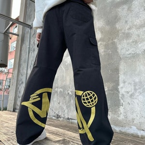 May include: Black cargo pants with a gold graphic design on each leg. The design features a circle with a stylized letter "W" and a globe inside the circle.