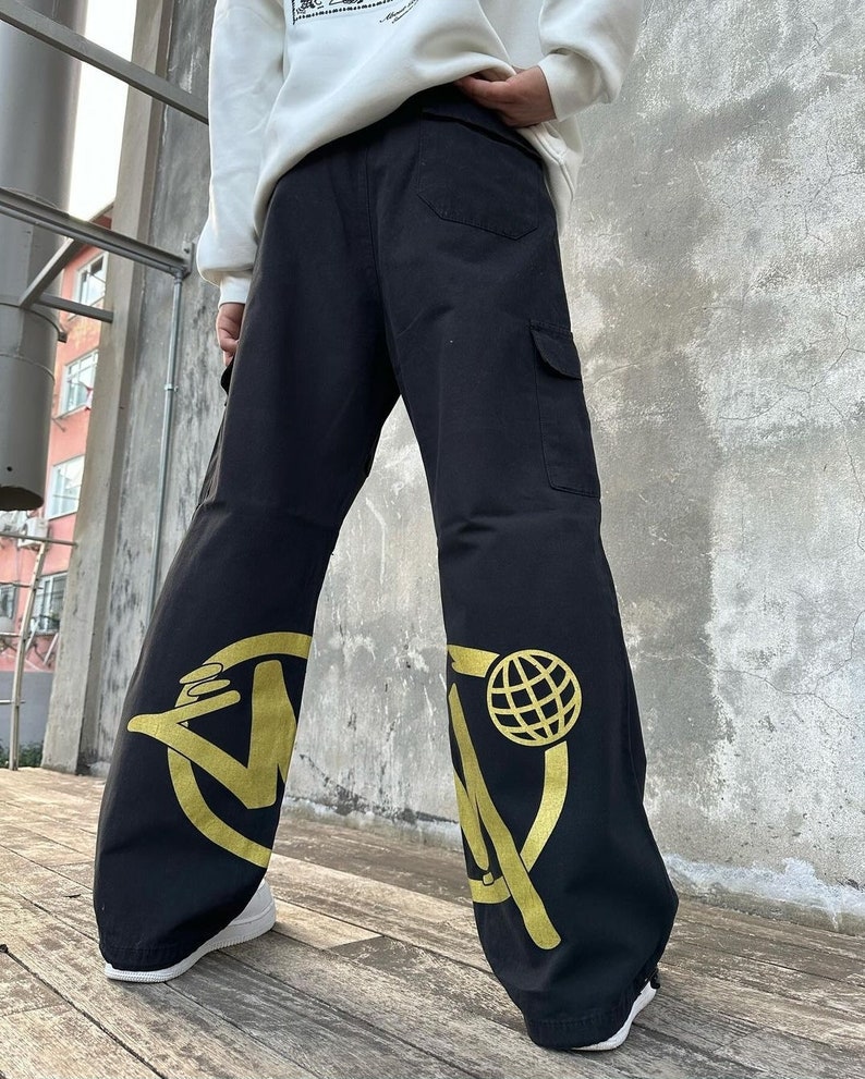May include: Black cargo pants with a gold graphic design on each leg. The design features a circle with a stylized letter "W" and a globe inside the circle.