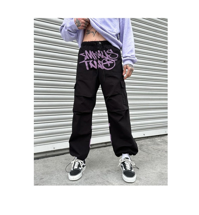 May include: Black cargo pants with a purple graphic print that says "MINUS TWO" on the front. The pants have a drawstring waist and multiple pockets.
