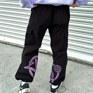 May include: Black cargo pants with purple graphic designs on the legs. The designs include a circle with a hand pointing out of it and a circle with a globe inside.