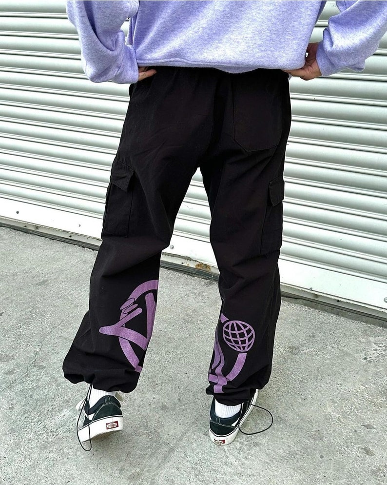 May include: Black cargo pants with purple graphic designs on the legs. The designs include a circle with a hand pointing out of it and a circle with a globe inside.
