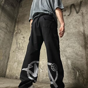 May include: Black cargo pants with a white graphic design on each leg. The design on the left leg features a hand holding a microphone, and the design on the right leg features a globe.