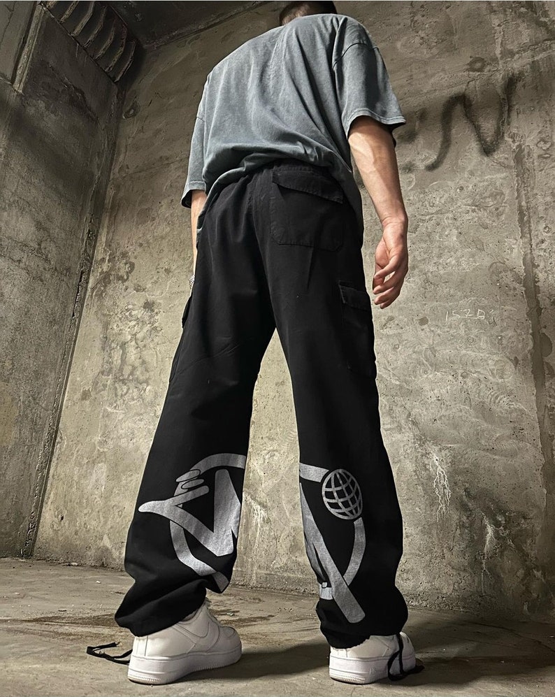 May include: Black cargo pants with a white graphic design on each leg. The design on the left leg features a hand holding a microphone, and the design on the right leg features a globe.