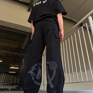 May include: Black cargo pants with a large graphic print of a circle with a globe and a stylized letter "M" on the left leg. The pants have a drawstring at the bottom and are worn with black sneakers.