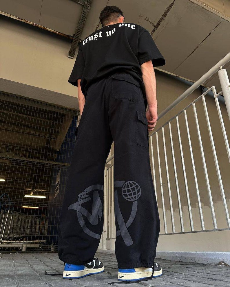 May include: Black cargo pants with a large graphic print of a circle with a globe and a stylized letter "M" on the left leg. The pants have a drawstring at the bottom and are worn with black sneakers.